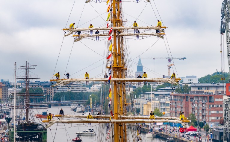 Kuvassa suuren purjealuksen masto, jossa miehistö kiipeilee, taustalla Turun kaupunkimaisemaa.